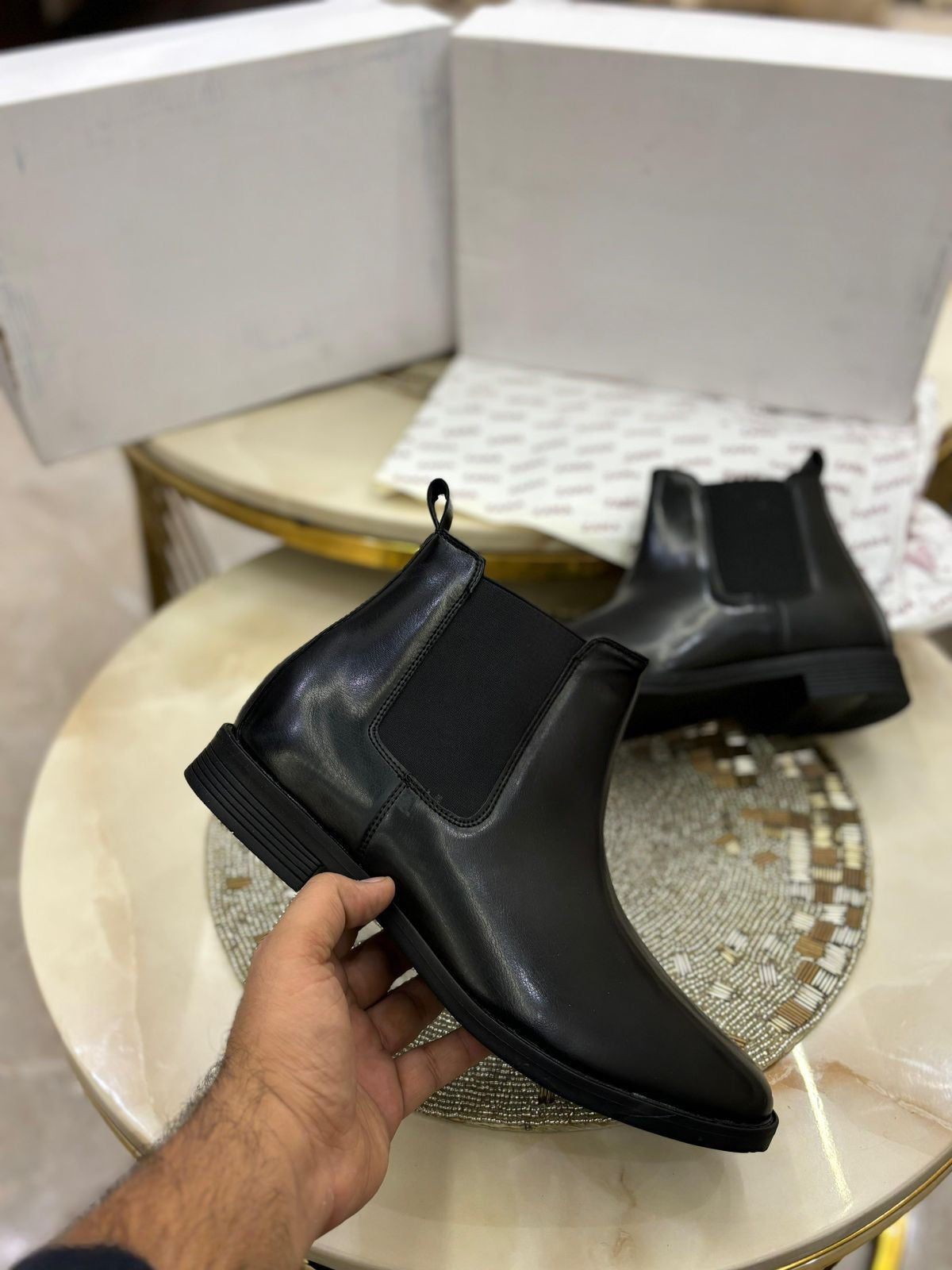 Black Chelsea boots held by a hand on a decorative table.