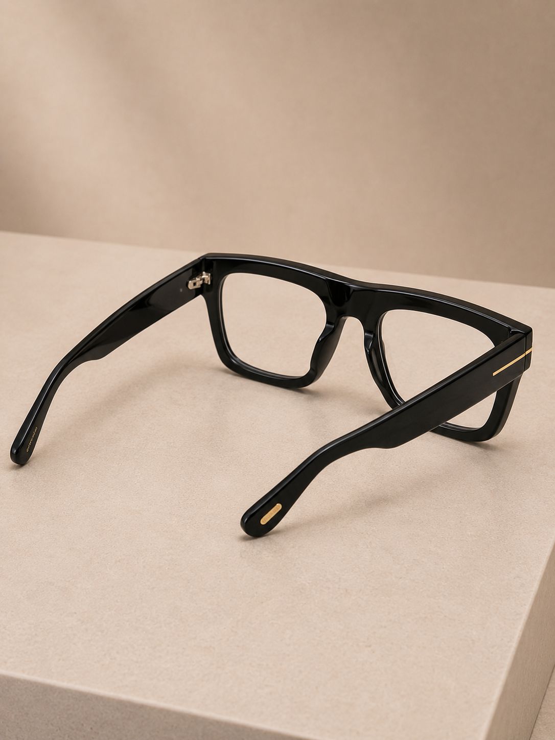 Black acetate square-frame eyeglasses with gold metal accents on the temples, resting on a beige surface against a matching beige background.