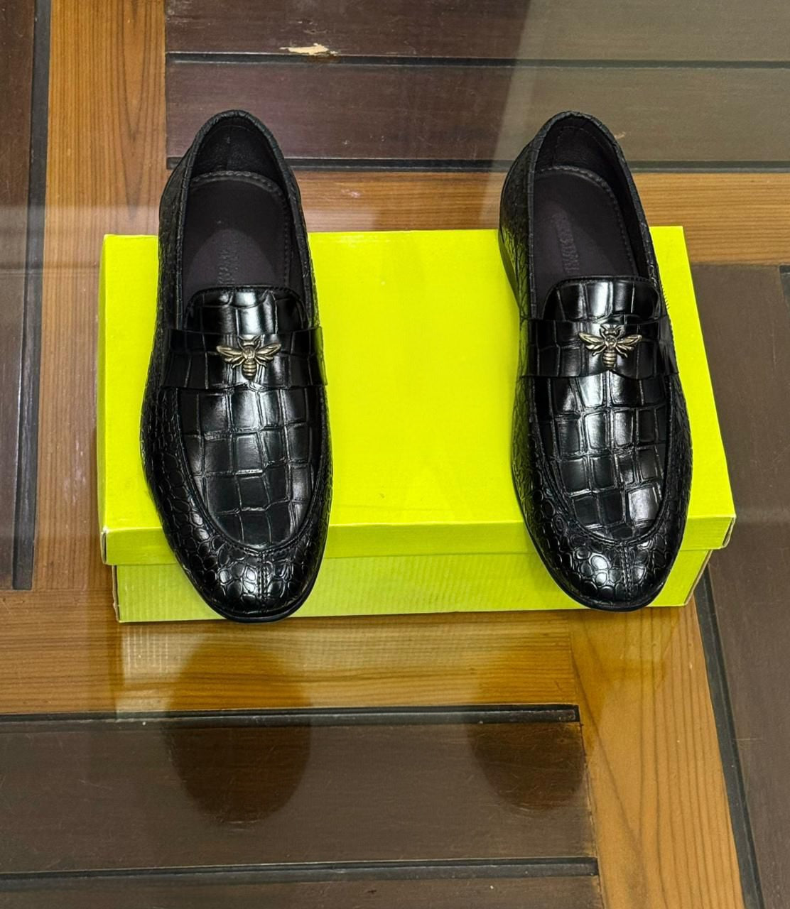 Black crocodile-patterned loafers on a yellow box with a wooden floor background