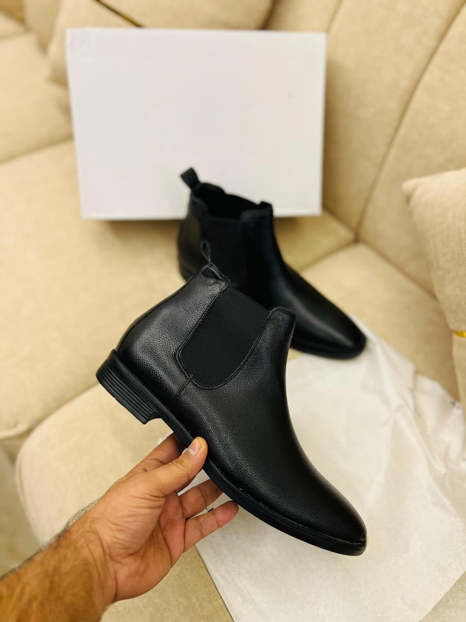 Black leather Chelsea boots being held on a beige couch with a white box in the background.