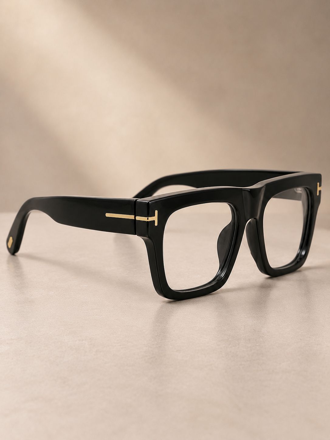 Black square-frame eyeglasses with gold metal accents on the temples, displayed on a neutral beige surface with soft shadows.