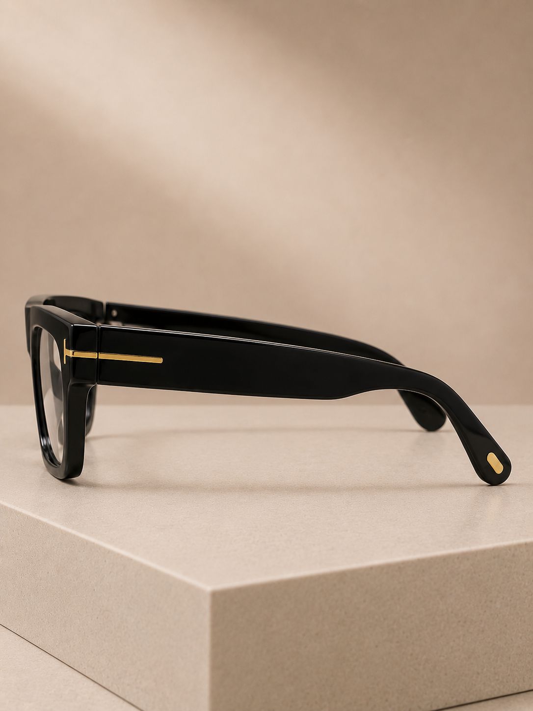Black square-frame eyeglasses with gold metal accents on the temples, resting on a beige display block against a beige background.