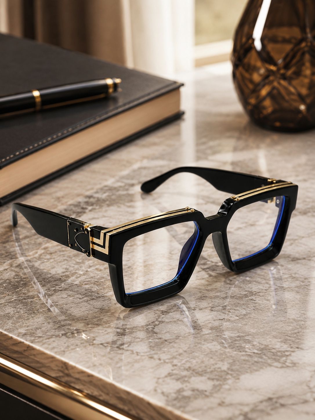 Black square eyeglasses with gold geometric trim and blue inner accents, resting on a marble desk next to a book and pen.