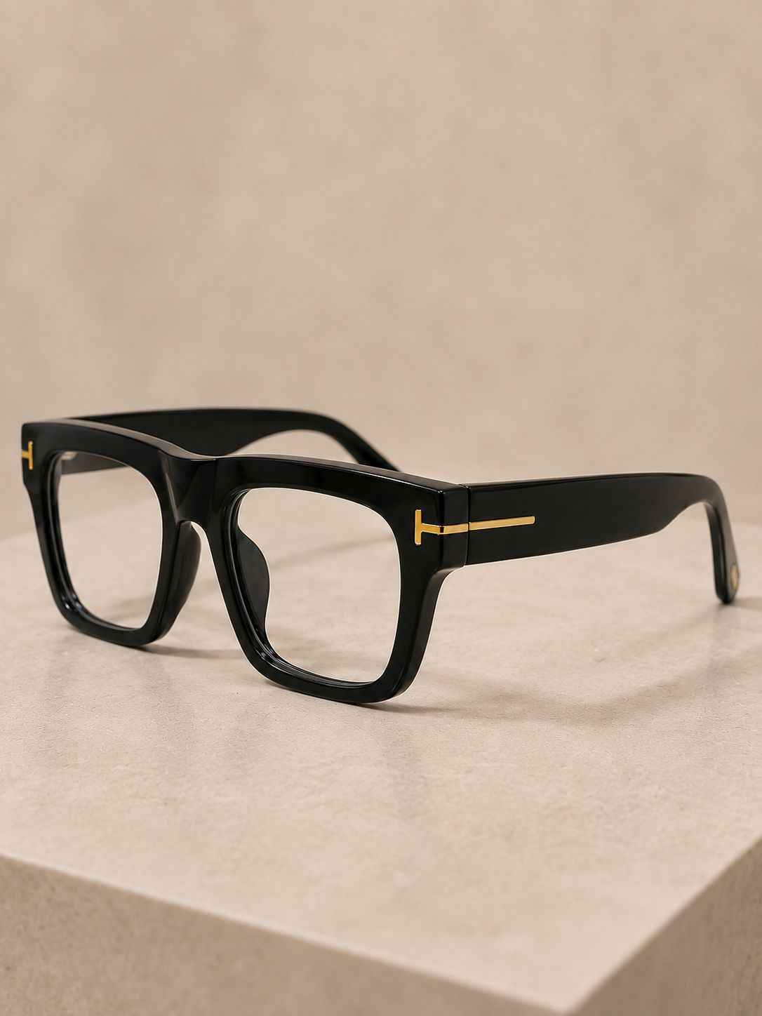 Black square eyeglasses with gold metal T-shaped accents on the temples, displayed on a light beige stone surface against a matching beige background.