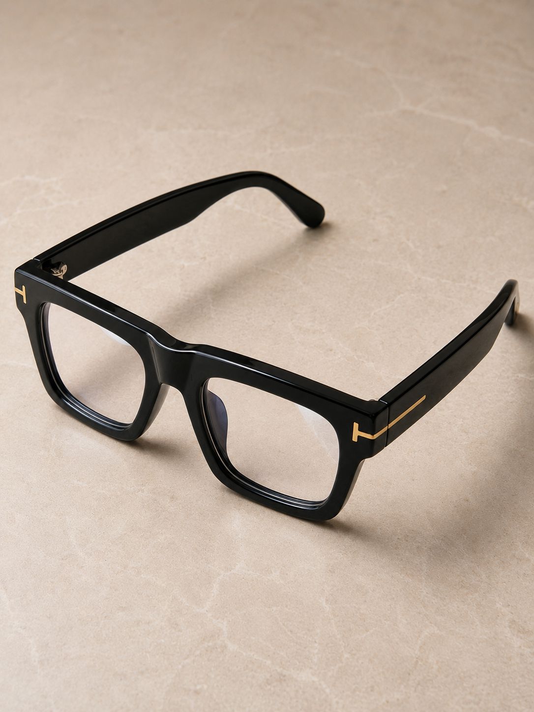 Black square eyeglasses with gold metal accents on the temples, displayed on a textured beige surface.