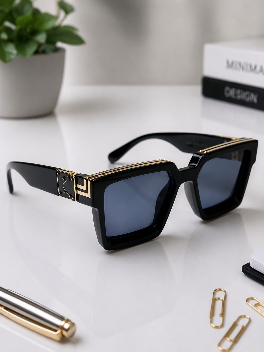 Black square sunglasses with gold geometric accents on the temples and dark blue-gray lenses, displayed on a white desk with a pen, paper clips, a plant, and books in the background.