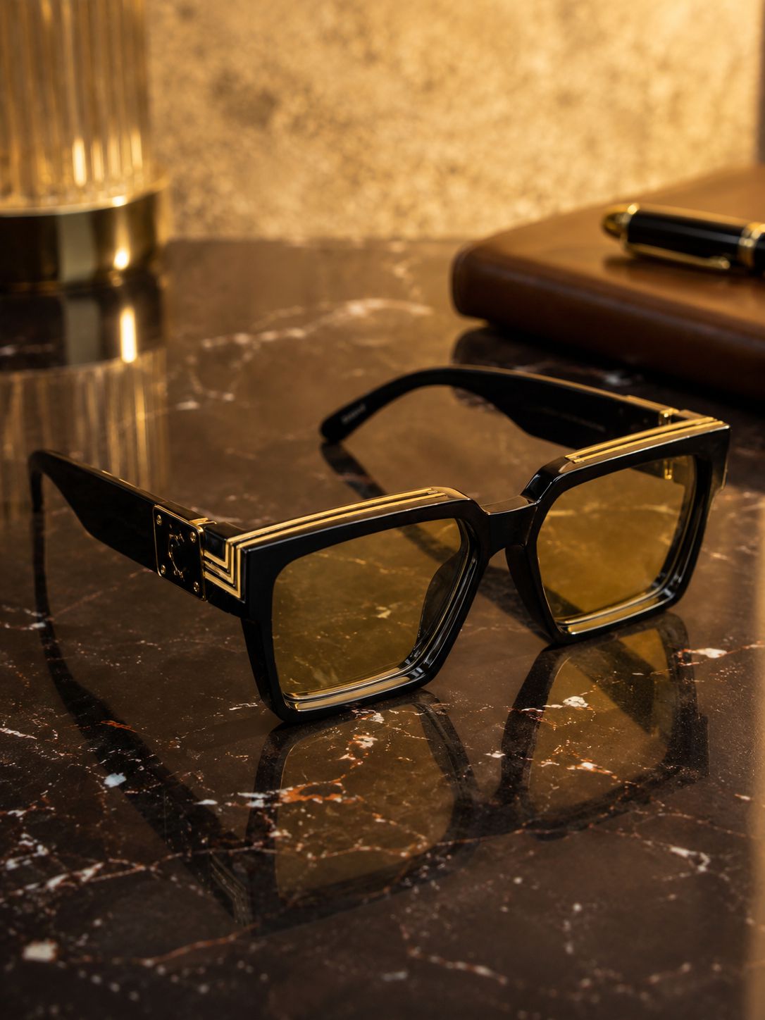 Black square sunglasses with gold geometric trim and yellow-tinted lenses, displayed on a dark marble surface with a warm, reflective background.
