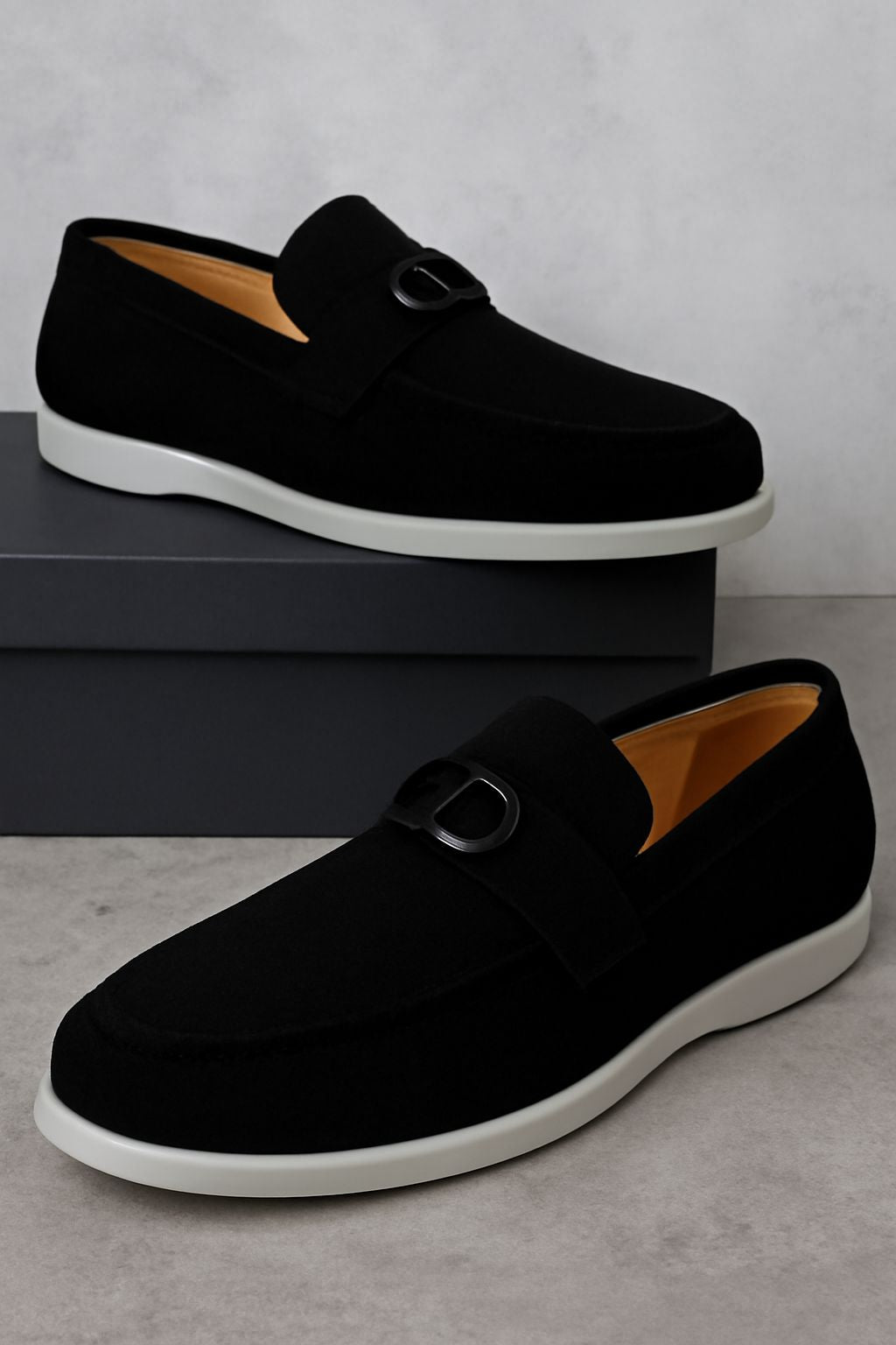 Black suede loafers with a sleek metal buckle and TPR sole resting on a gray shoebox.