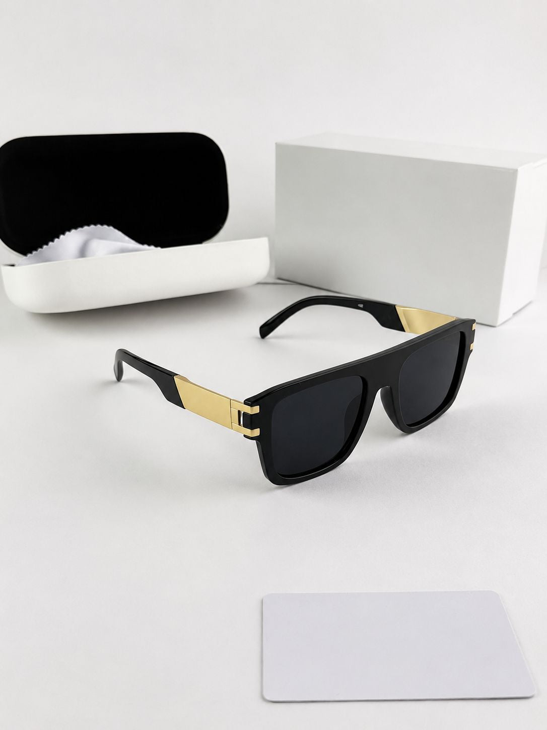 Black sunglasses with gold accents on a white surface, accompanied by a white case and cleaning cloth.