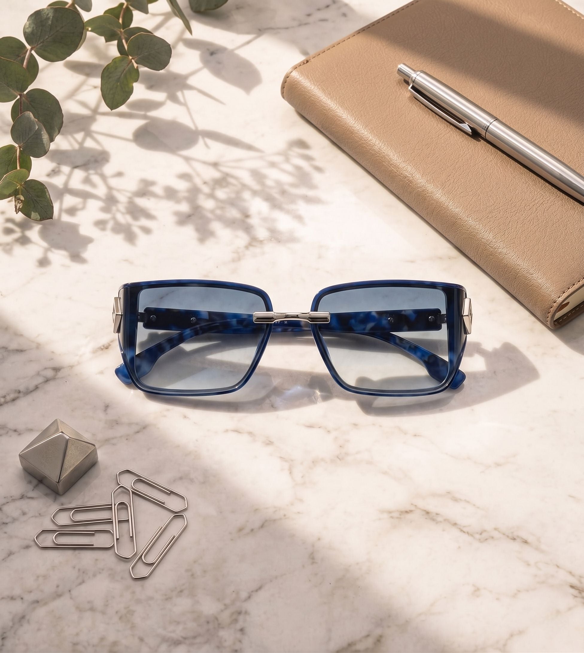 Blue sunglasses on a marble surface with a pen, notebook, and decorative items.