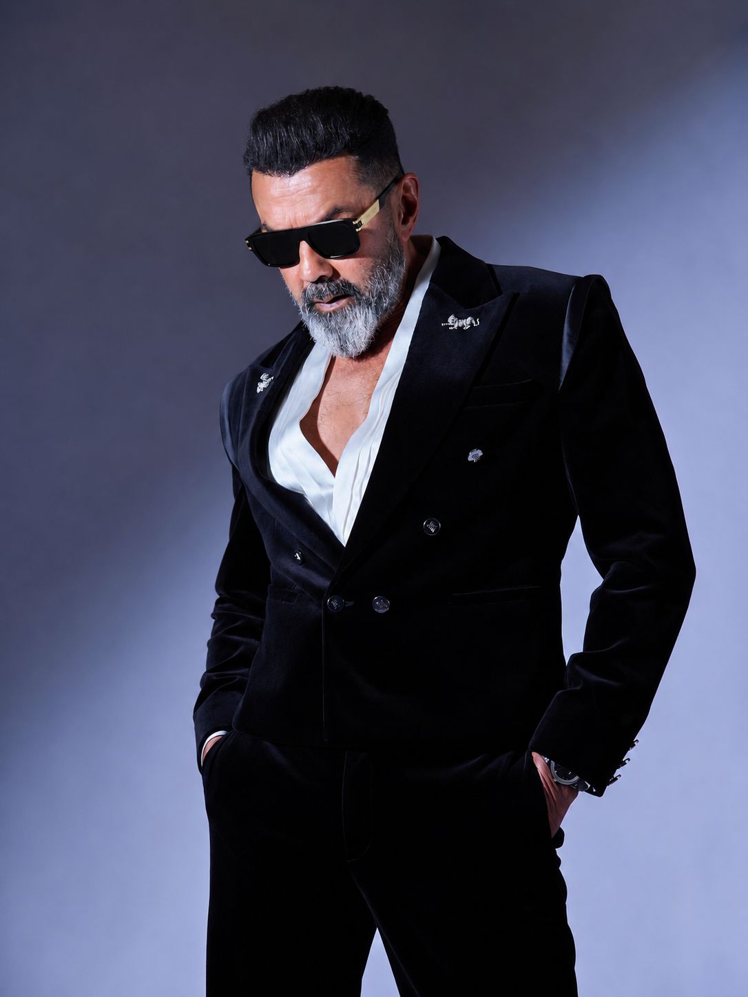 Bobby deol wearing a black suit with sunglasses and a gray beard against a gray background