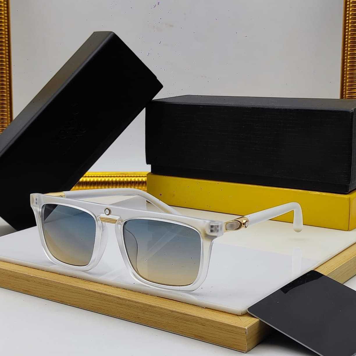 Clear white rectangular sunglasses with gold accents and blue to yellow gradient lenses, displayed on a marble block with black boxes.