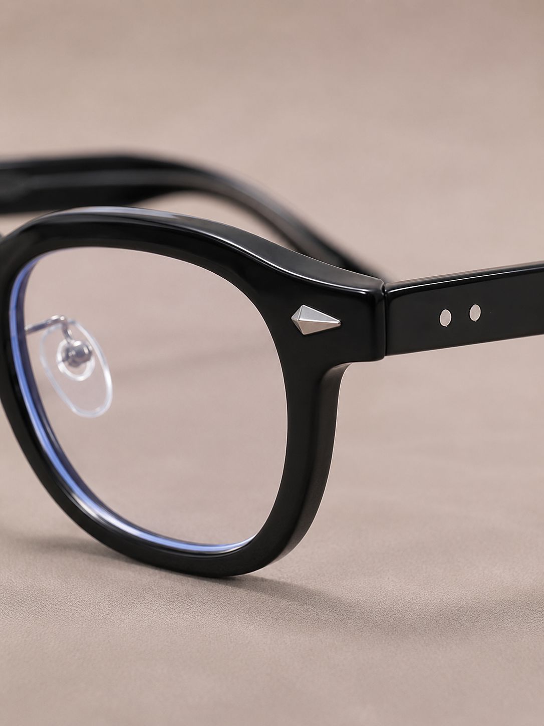 Close-up of a pair of black acetate eyeglass frames with a geometric triangular metal accent on the hinge, resting on a neutral beige surface.