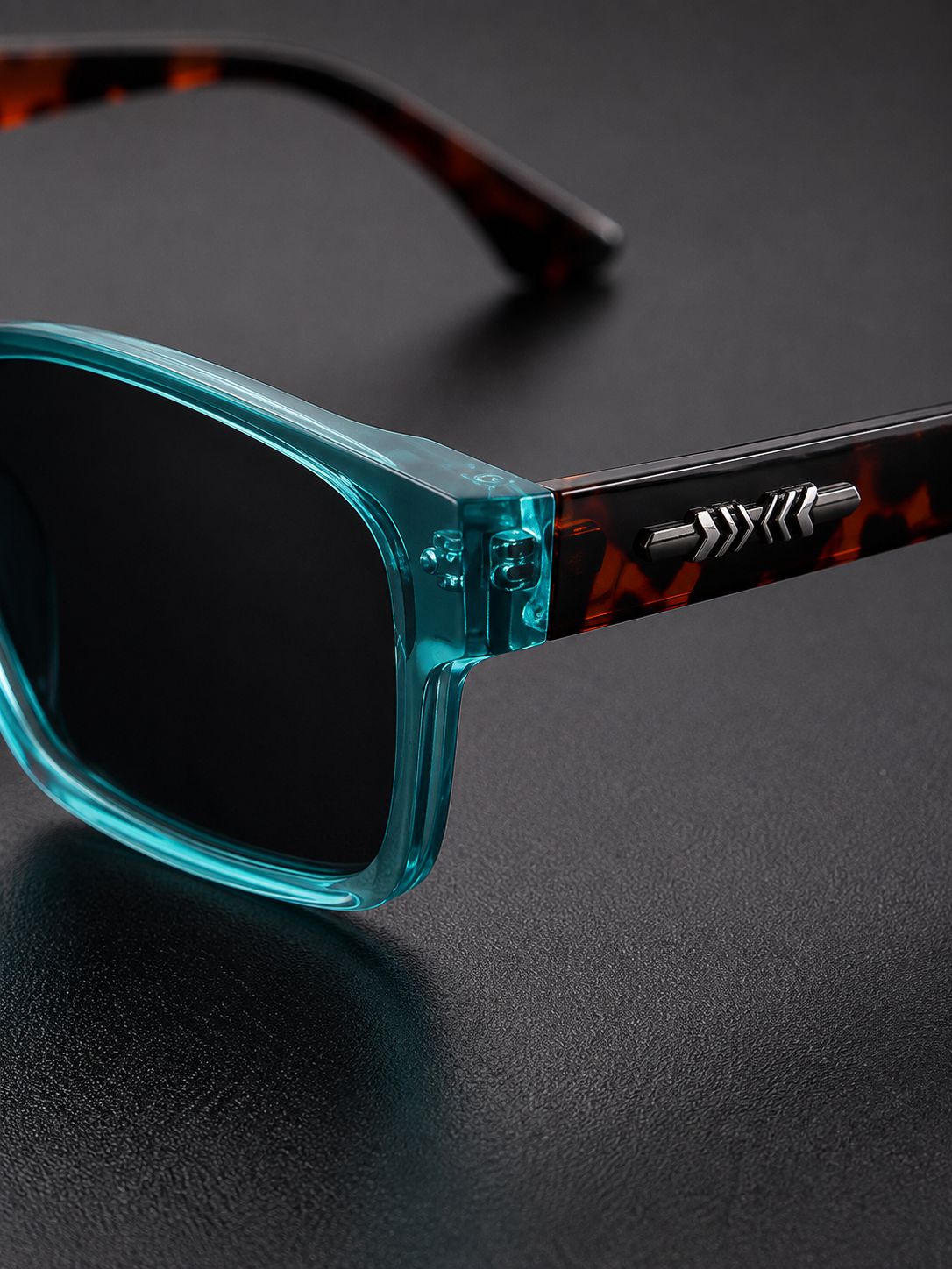 Close-up of a pair of sunglasses with translucent teal-blue front frames and dark tortoiseshell temples, resting on a dark textured surface.