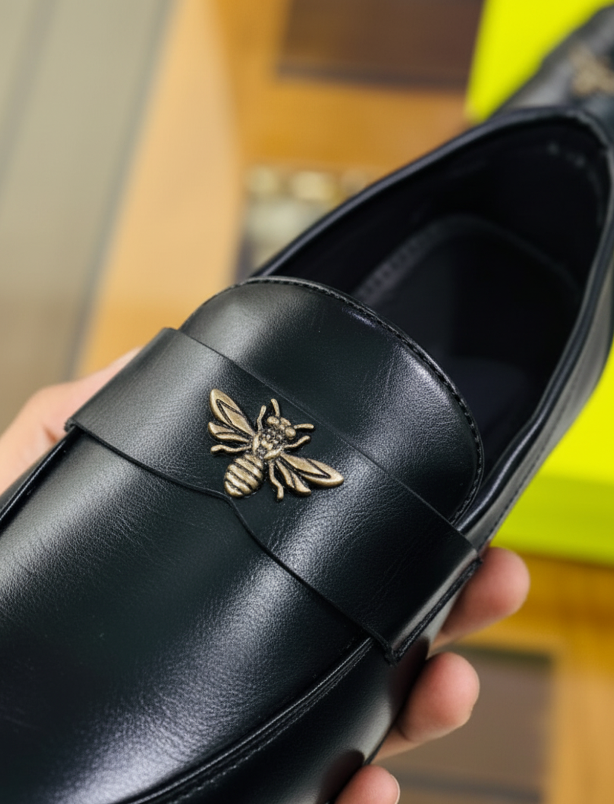 Close-up of gold bee emblem on black faux leather loafer, held in hand