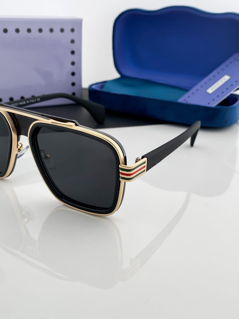 Close-up of the square aviator sunglasses showing the gold metal frame and the distinctive green-red stripe pattern on the temple.