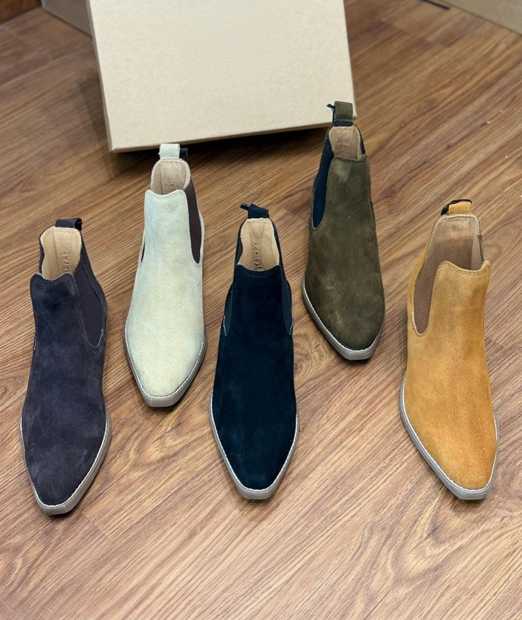 Five pairs of suede ankle boots in different colors on a wooden floor.