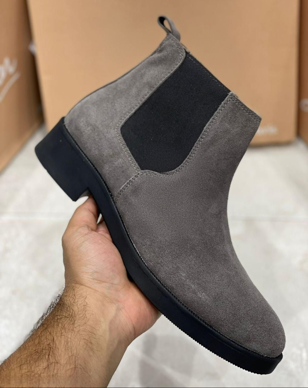 Gray suede Chelsea boot held by a hand with a cardboard box in the background