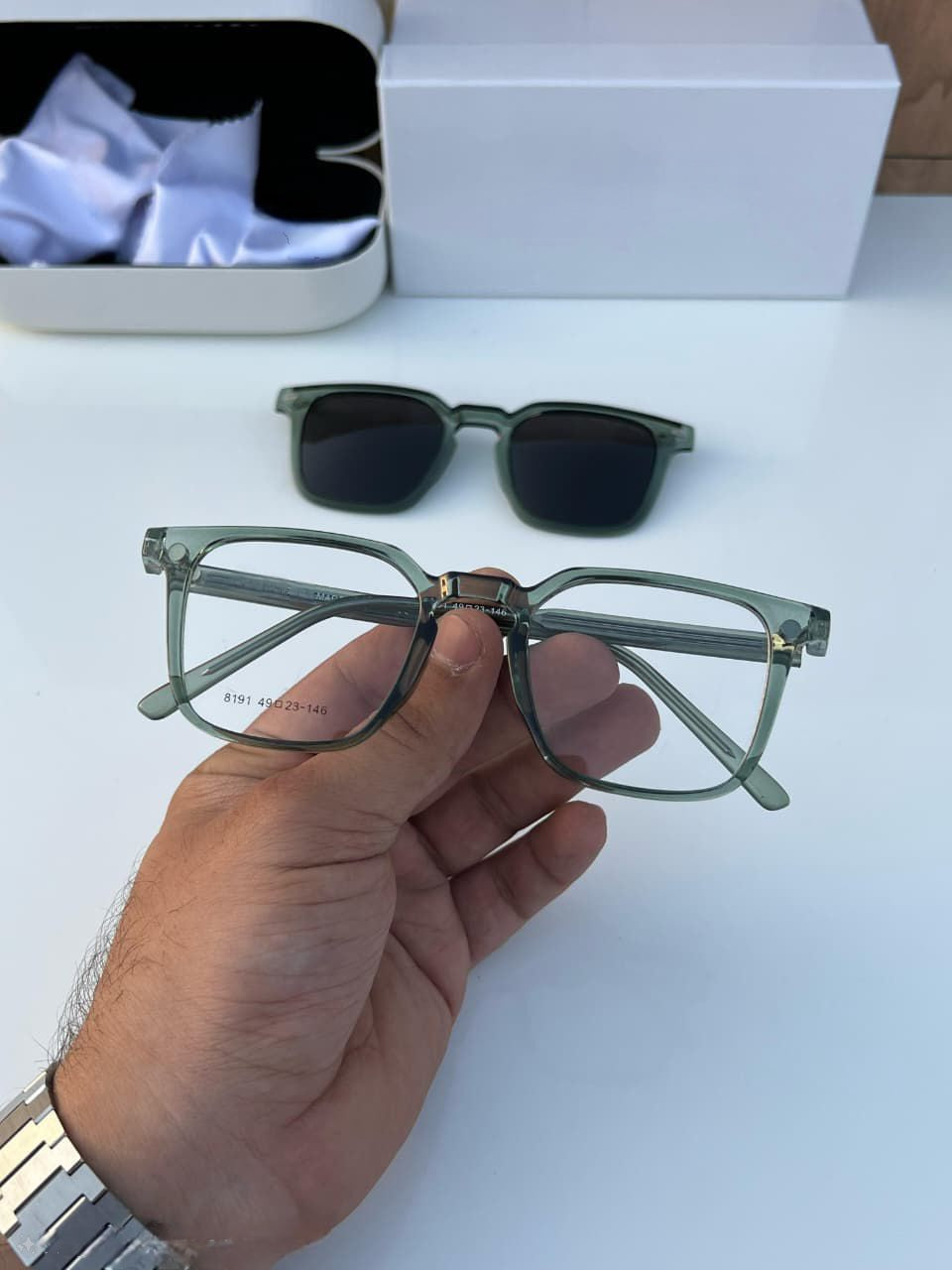 Hand holding the Transparent Grey/Green Base Frame Eyeglasses with the dark clip-on lying behind.