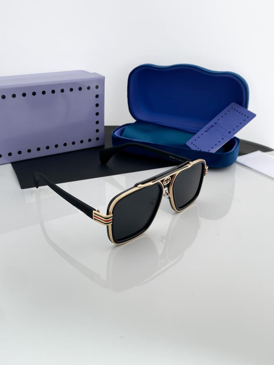 Luxury square aviator sunglasses with a gold metal frame, black lenses, and green-red stripe detail on the temples, displayed with blue and purple packaging.