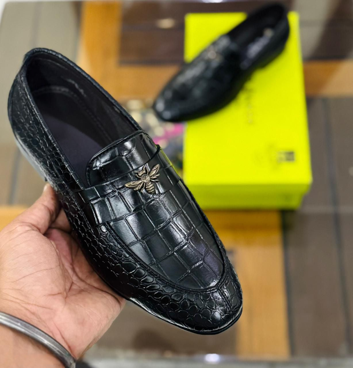 Man holding a black crocodile-embossed loafer with a gold bee, showing the texture and design clearly.