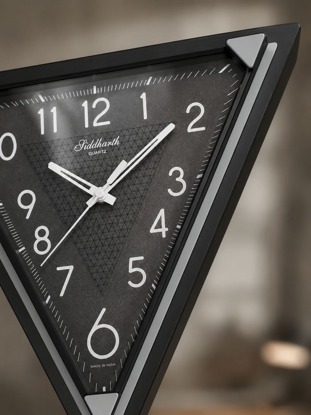 A close-up of a triangular wall clock with a black frame and textured dark gray dial, featuring white Arabic numerals and hands. The brand name 'Siddharth' is visible on the dial.