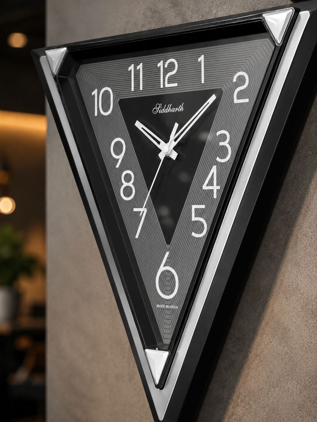 A triangular wall clock with a black frame and textured dark gray face, featuring white Arabic numerals and hands. The brand name 'Feldharte' is visible in script near the center.
