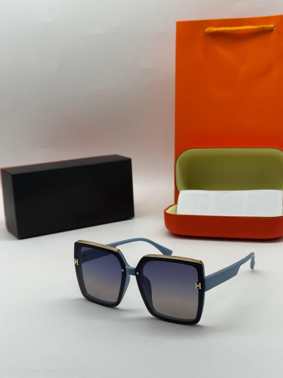 Oversized light blue square sunglasses with gold trim and H-logo detail and blue-grey gradient lenses.