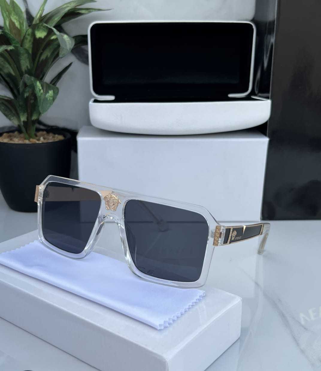 Transparent square frame sunglasses with dark lenses and gold Medusa-like emblem on temple.