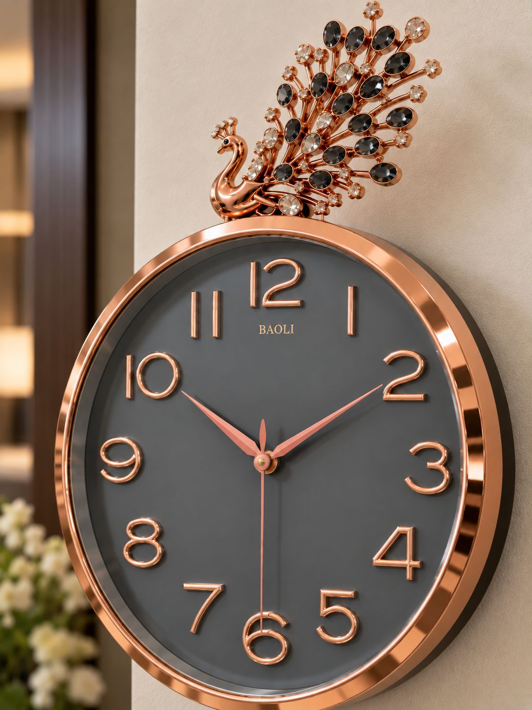 round wall clock with a rose gold frame, dark gray dial, and a decorative peacock topper with black rhinestones, shown mounted on a beige wall.