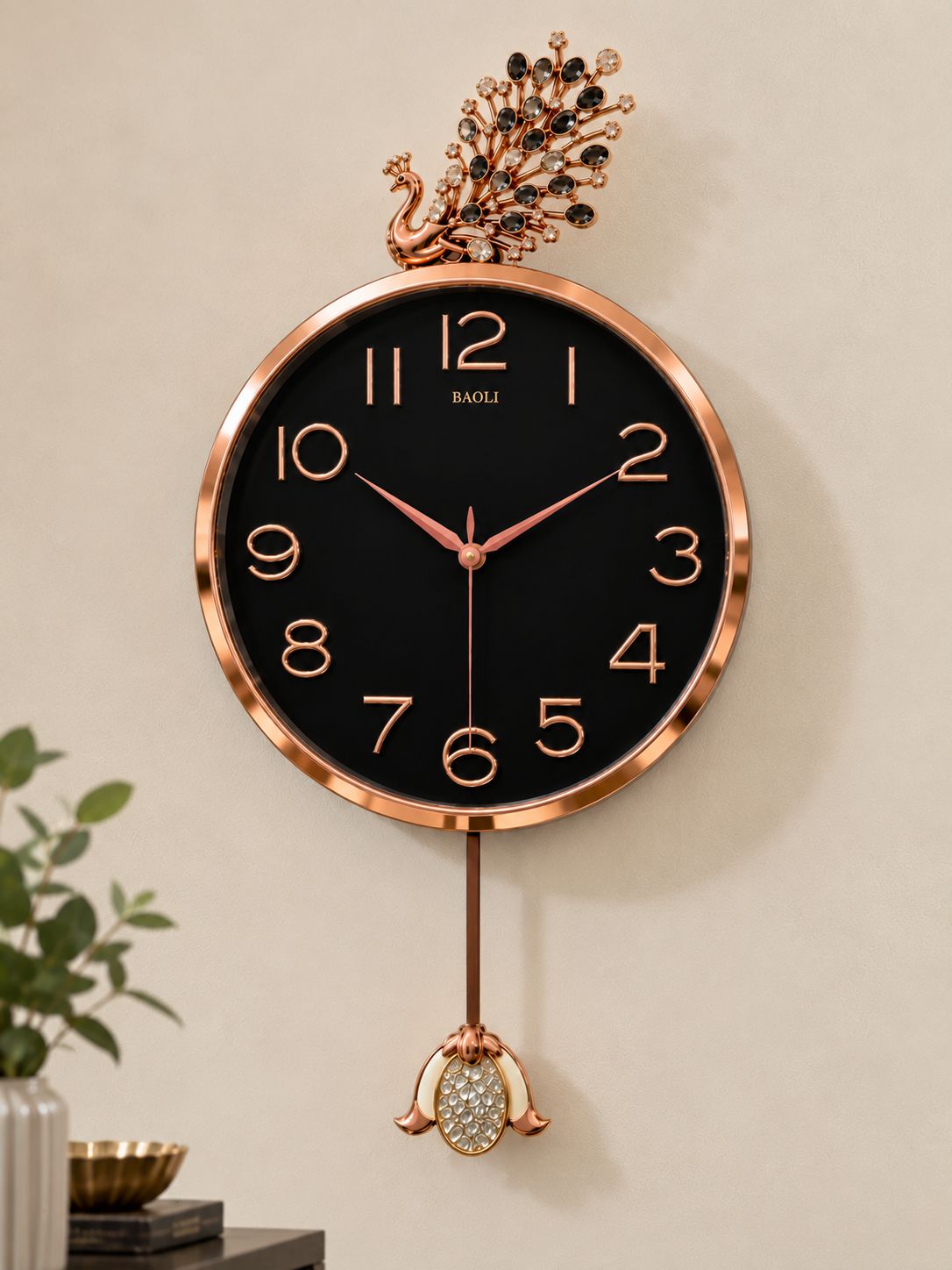wall clock with a peacock top and hanging ornament, featuring a black dial and rose gold frame, shown mounted on a beige wall.